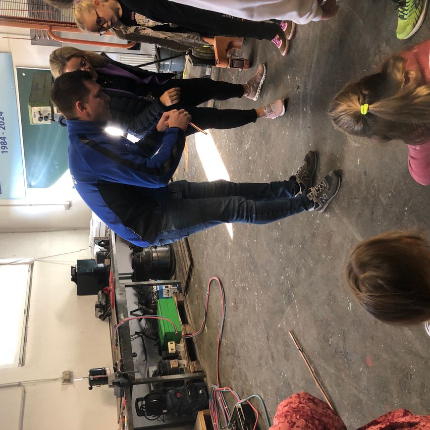 Eine Gruppe von Menschen befindet sich in einer Werkstatt. Ein Mann ist gebeugt und hält einen Bleistift. Mehrere Kinder beobachten. Geräte stehen auf einem Tisch, und Kabel liegen auf dem Boden verstreut.