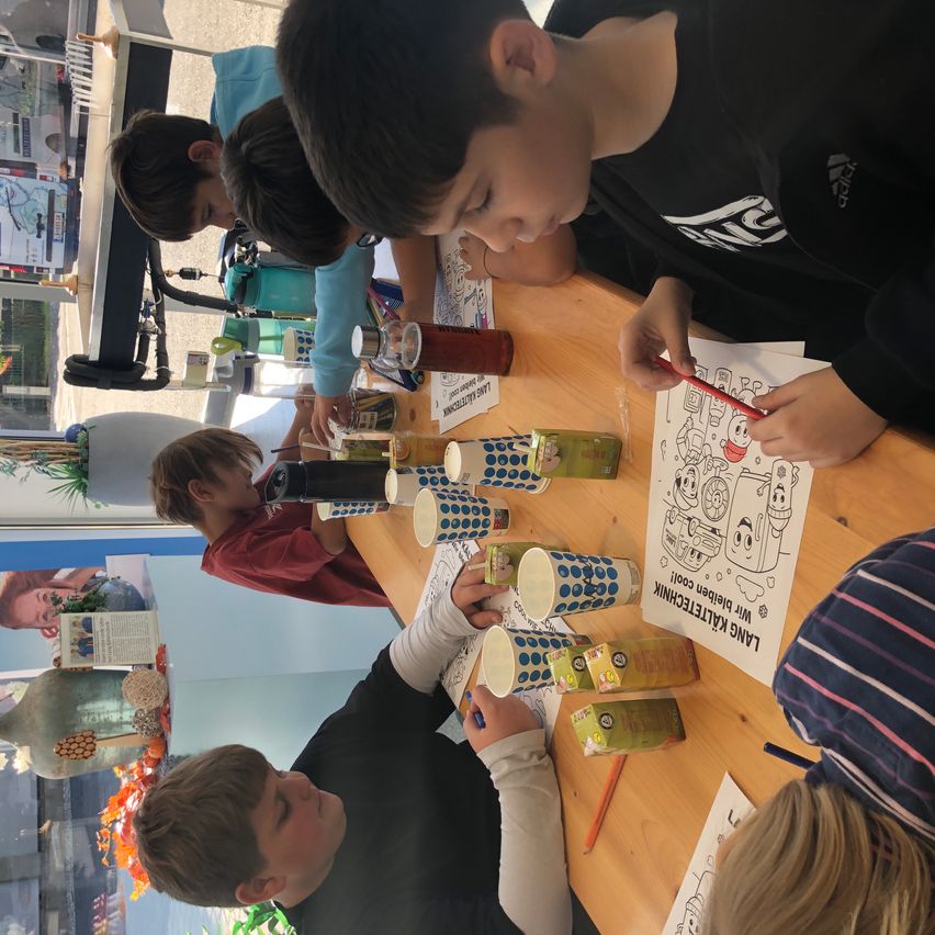 Eine Gruppe von Kindern sitzt an einem Tisch und malt ein Bild mit Markern und Bleistiften. Verschiedene Gegenstände sind auf dem Tisch, einschließlich Becher und Flaschen. Die Kinder konzentrieren sich auf ihre Aktivität.