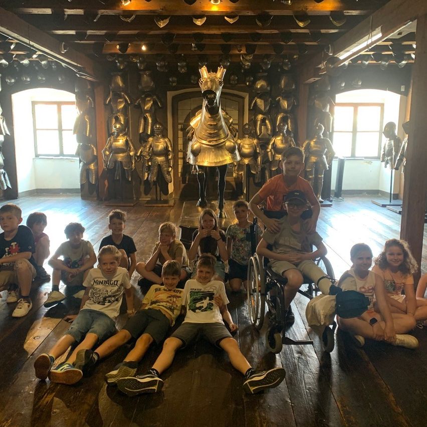 Eine Gruppe von Kindern und einem jungen Mann sitzen auf dem Boden eines Raumes. Hinter ihnen stehen goldene Statuen von Rittern und einem Pferd.