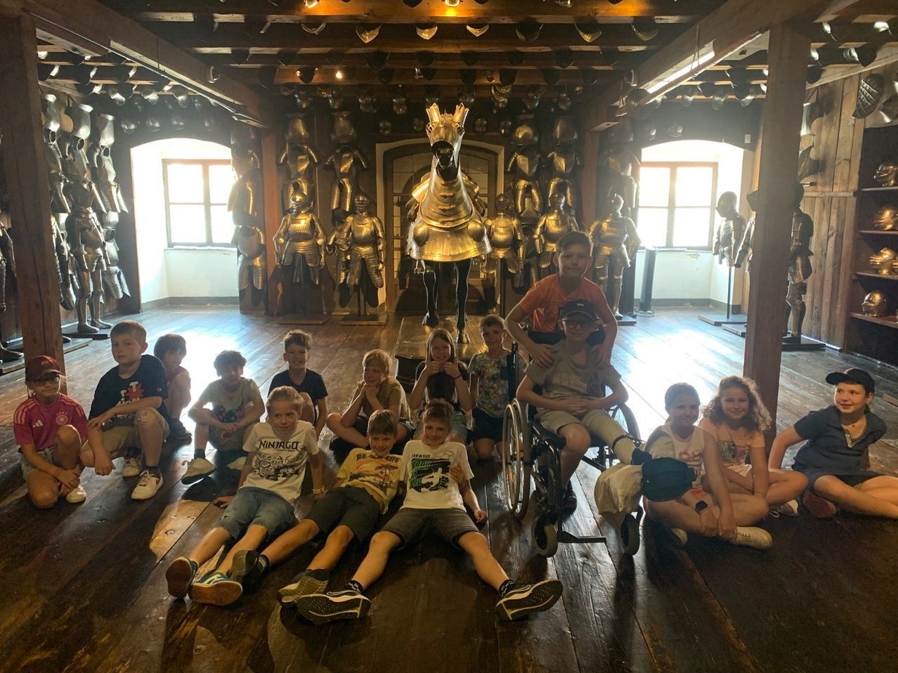 Eine Gruppe von Kindern und einem jungen Mann sitzen auf dem Boden eines Raumes. Hinter ihnen stehen goldene Statuen von Rittern und einem Pferd.