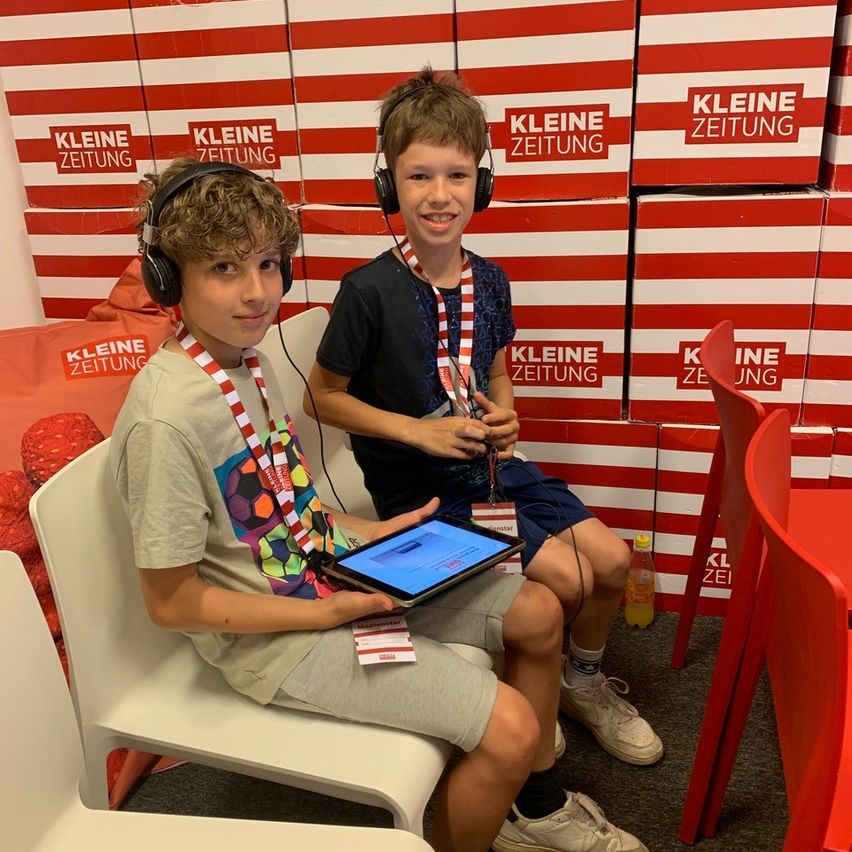 Zwei Jungen sitzen auf Stühlen und tragen Kopfhörer. Einer hält ein Tablet. Sie stehen vor Kisten mit Kleine Zeitung darauf geschrieben.