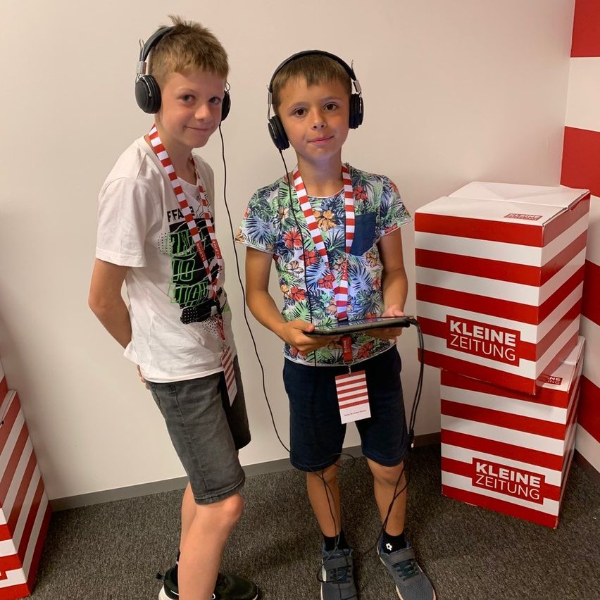 Zwei Jungen mit Kopfhörern, die neben Schachteln mit der Aufschrift Kleine Zeitung stehen, tragen lässige Sommerkleidung und scheinen etwas auf einem Tablet zu hören.