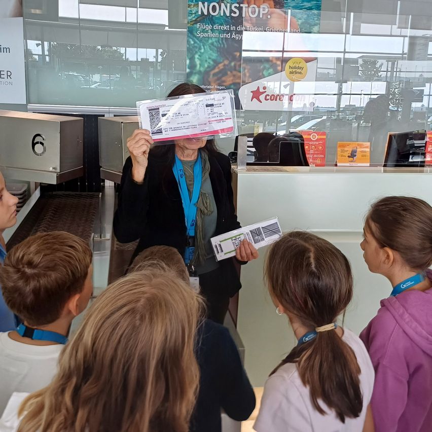 Eine Frau mit Reisepass und Tickets steht in einem Flughafen, umgeben von einer Gruppe von Kindern, die sie anschauen.