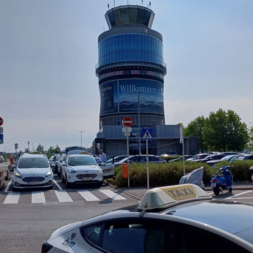 Ein geschäftiger Flughafenparkplatz mit aufgereihten Taxis, einem Kontrollturm und einer großen Mercedes-Benz-Werbung.