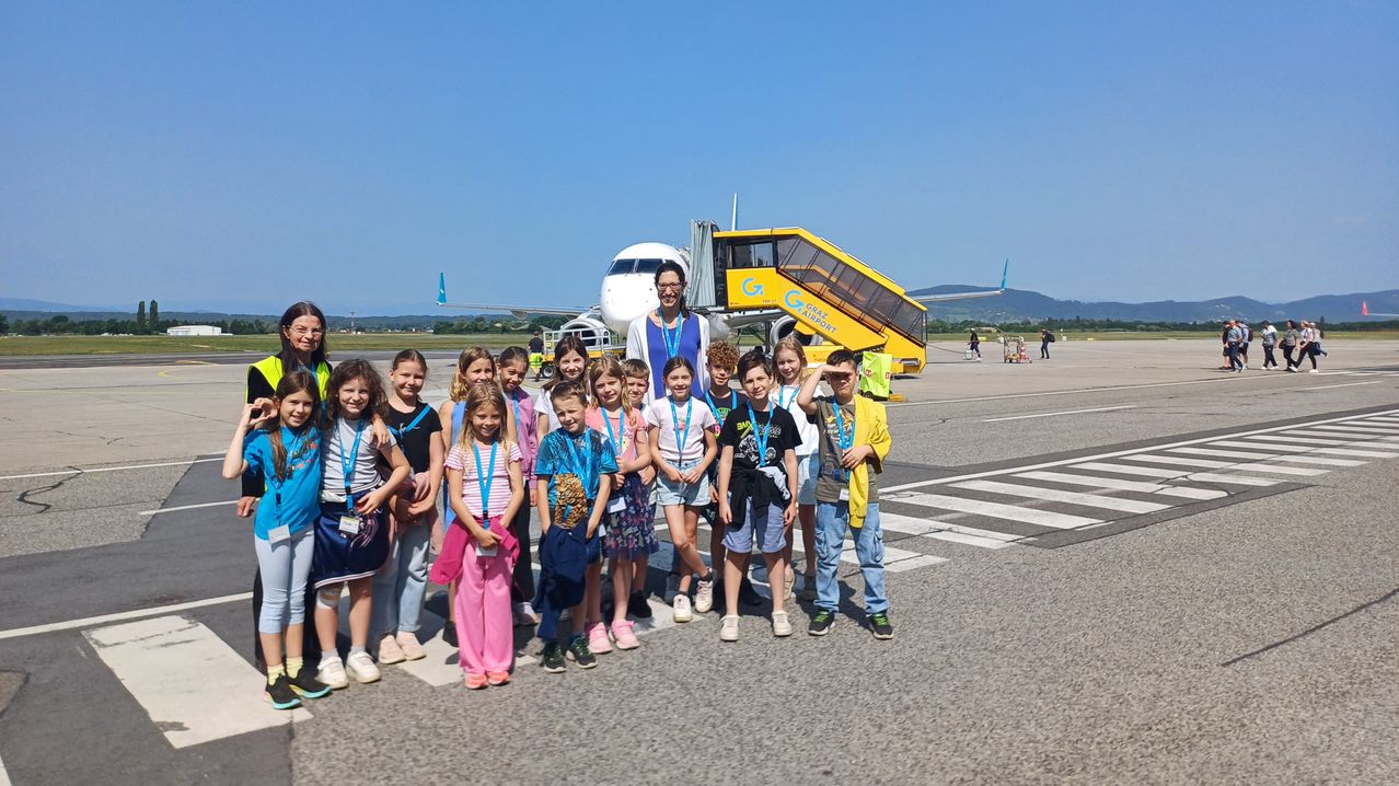 Eine Gruppe von Kindern mit Namensschildern steht auf dem Vorfeld eines Flughafens und posiert für ein Foto mit einem Flugzeug und einem gelben Bus im Hintergrund.