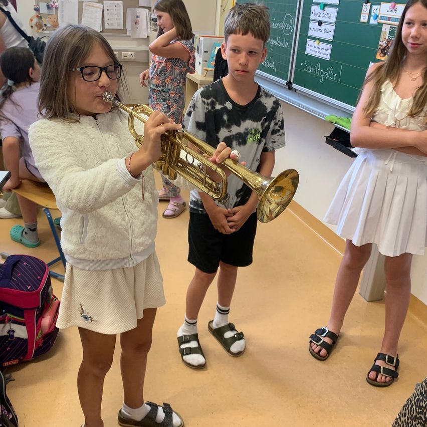 Ein Mädchen spielt Trompete in einem Klassenzimmer, während ein Junge zusieht. Ein anderes Mädchen beobachtet sie. Hinter ihnen steht eine Tafel mit Schriften und Zeichnungen.