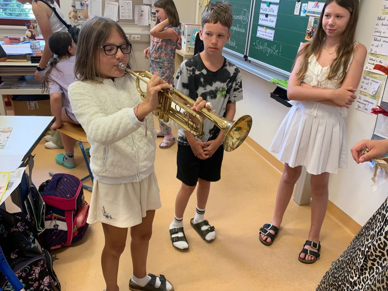 Ein Mädchen spielt Trompete in einem Klassenzimmer, während ein Junge zusieht. Ein anderes Mädchen beobachtet sie. Hinter ihnen steht eine Tafel mit Schriften und Zeichnungen.