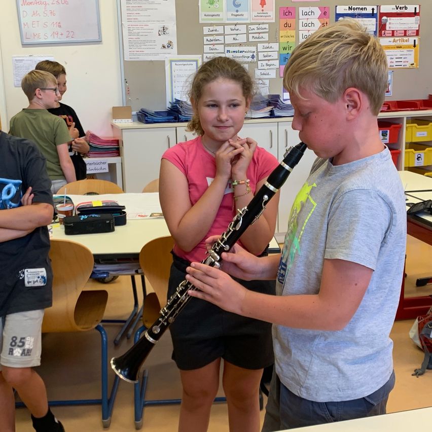 In einem Klassenzimmer spielen ein junges Mädchen in einem rosa T-Shirt und ein Junge in einem grauen T-Shirt gemeinsam eine Klarinette. Hinter ihnen beobachten andere Kinder, und es gibt Schreibtische und eine Pinnwand mit verschiedenen angehefteten Papieren.