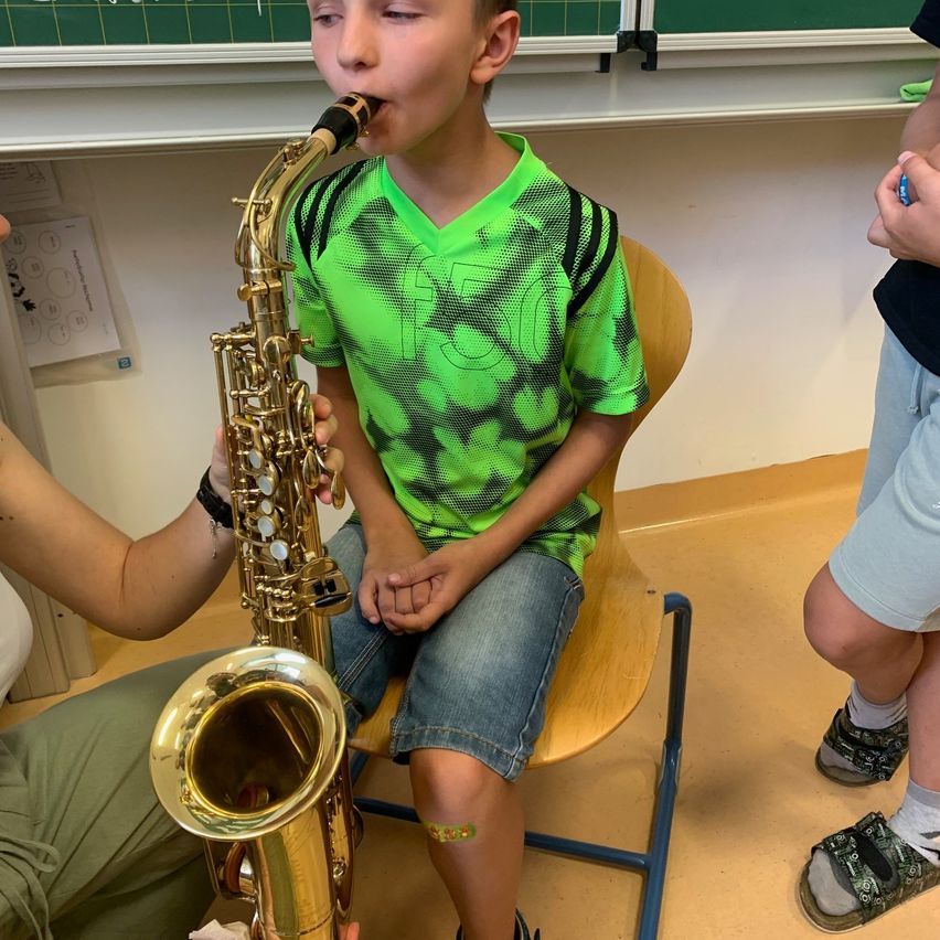 Ein junger Junge in einem grünen Trikot spielt Saxophon, sitzt in einem Klassenzimmer, während eine Person einen Stift hält und eine andere zusieht.