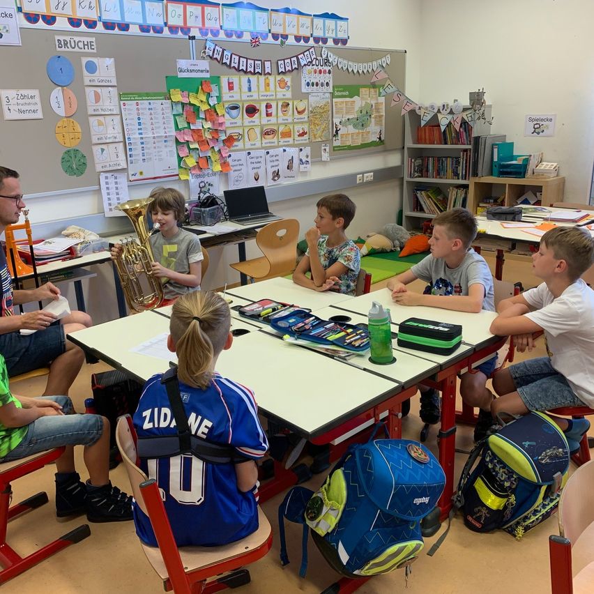 Eine Gruppe von Kindern und einem Erwachsenen befindet sich in einem Klassenzimmer. Die Kinder sitzen um einen Tisch herum, während der Erwachsene Tuba spielt. Das Klassenzimmer ist mit verschiedenen Postern und Büchern dekoriert.