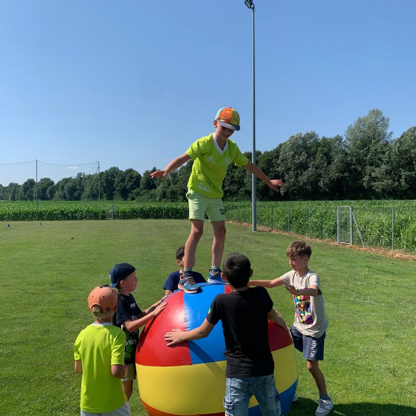Eine Gruppe von Kindern spielt mit einem großen, mehrfarbigen Ball auf einem Rasenfeld unter klarem Himmel.
