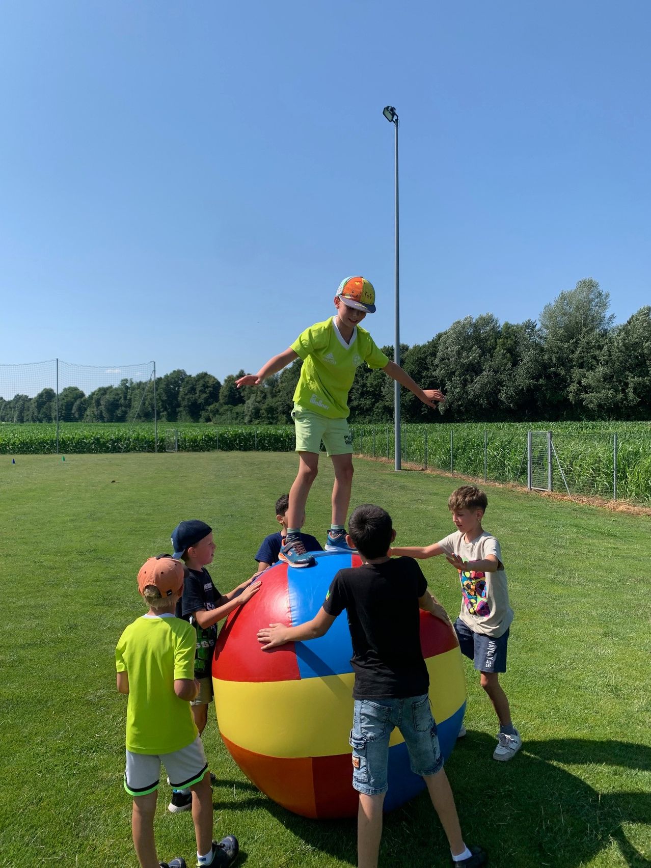 Eine Gruppe von Kindern spielt mit einem großen, mehrfarbigen Ball auf einem Rasenfeld unter klarem Himmel.