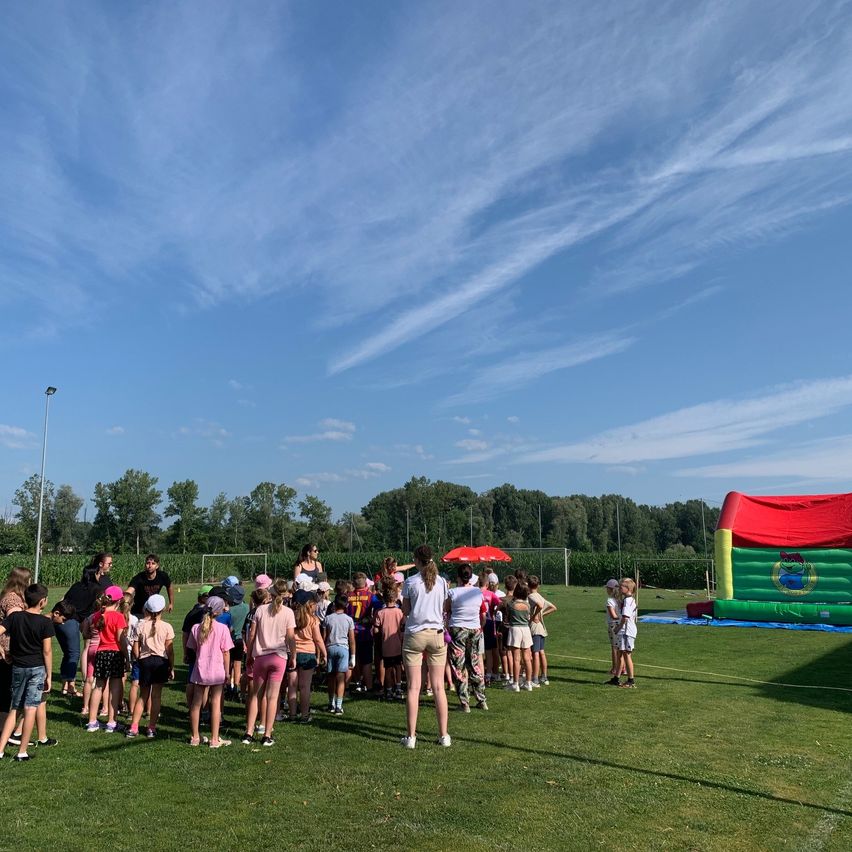 Eine Gruppe von Kindern und Erwachsenen steht auf einem Rasenfeld mit einem aufblasbaren Schloss. Einige tragen Hüte und Turnschuhe. Eine Frau hält einen Regenschirm in der Nähe eines Mannes in einem schwarzen Shirt. Bäume und eine Straßenlaterne sind in der Ferne.