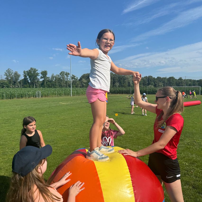 Ein Mädchen steht auf einem großen aufblasbaren Ball und hält die Hände einer Frau. Andere Kinder beobachten es auf einer Wiese.