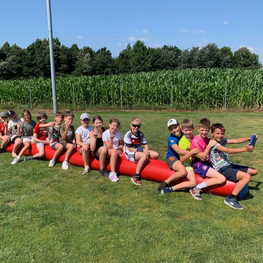 Eine Gruppe von Kindern sitzt auf einer roten aufblasbaren Röhre in einem Grasfeld und posiert für ein Foto mit einem Maisfeld im Hintergrund.