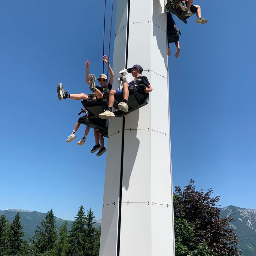 Drei Personen fahren auf einer Turmfahrt. Sie tragen Mützen und Turnschuhe. Dahinter steht ein weißer Turm mit Bergen im Hintergrund.