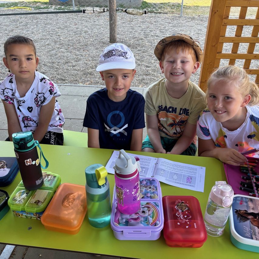 Vier Kinder sitzen auf einer Bank im Freien. Sie lächeln und scheinen für ein Foto zu posieren. Vor ihnen steht ein grüner Tisch mit verschiedenen Gegenständen wie Brotdosen, Wasserflaschen und Büchern.