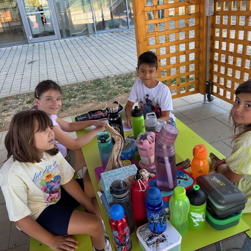 Vier Kinder sitzen um einen grünen Tisch herum, auf dem verschiedene Gegenstände wie Wasserflaschen, ein Lunchbox und ein Buch stehen. Die Kinder lächeln und scheinen ihre Mahlzeit zu genießen. Im Hintergrund ist eine Holzstruktur und ein Gebäude mit Glasfenstern zu sehen.