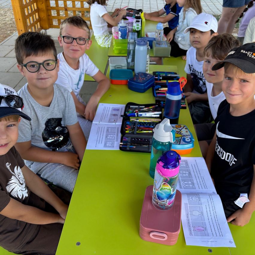 Fünf Jungen sitzen an einem grünen Picknicktisch mit verschiedenen Gegenständen. Einer trägt eine Brille, ein anderer einen Baseballcap, und es gibt Flaschen, Papiere und eine Brotdose.