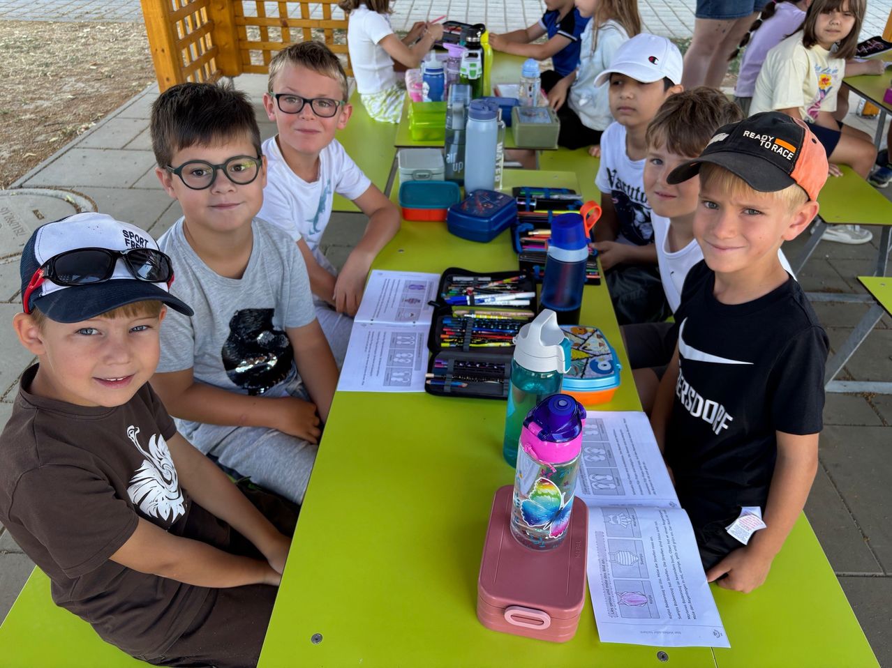 Fünf Jungen sitzen an einem grünen Picknicktisch mit verschiedenen Gegenständen. Einer trägt eine Brille, ein anderer einen Baseballcap, und es gibt Flaschen, Papiere und eine Brotdose.