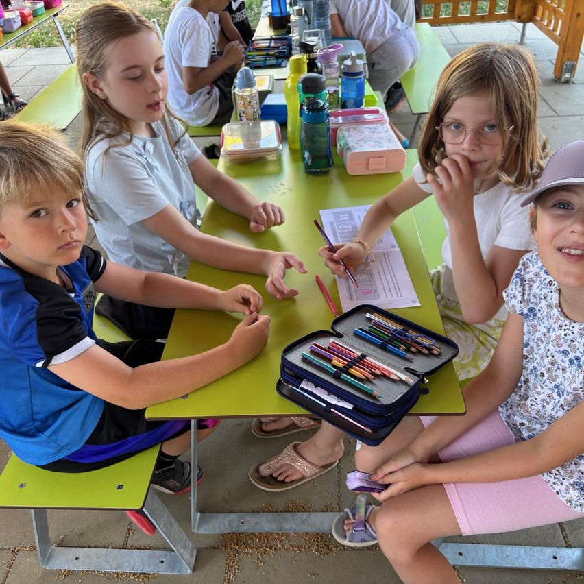 Vier Kinder sitzen an einem grünen Picknicktisch, zwei Mädchen und zwei Jungen, die sich mit Buntstiften und Papieren beschäftigen.