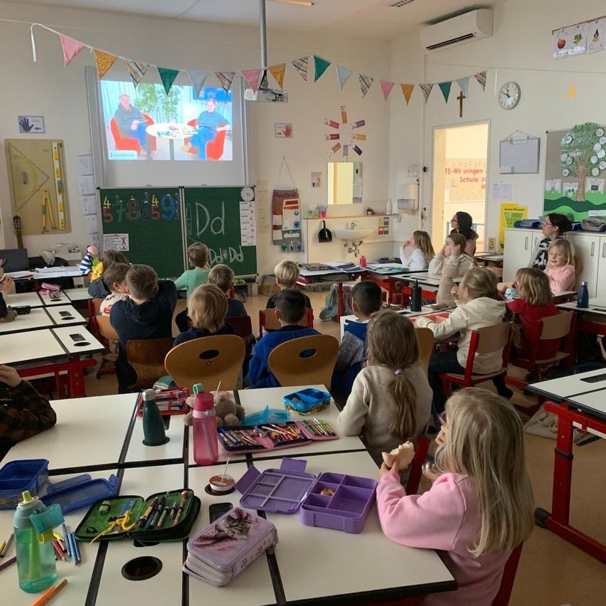 Ein Klassenzimmer mit Kindern an Schreibtischen, die eine Präsentation auf einer Projektionsleinwand ansehen. Bunte Dekorationen und pädagogische Poster schmücken die Wände.
