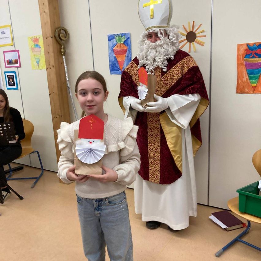 Ein junges Mädchen, das eine papierne Weihnachtsmanndekoration in der Hand hält, steht vor einem Mann, der als Sankt Nikolaus verkleidet ist, in einem Klassenzimmer. Hinter ihnen befindet sich eine Wand mit verschiedenen Kunstwerken.