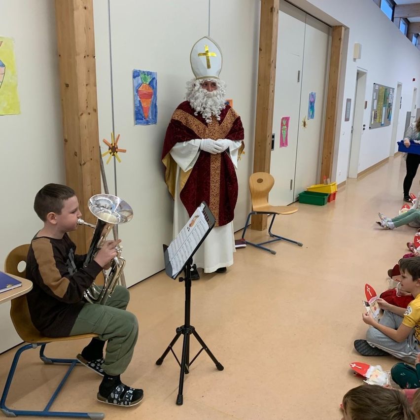 Ein Junge spielt die Trompete, während er eine grüne Uniform trägt. Hinter ihm steht ein Mann, der als Weihnachtsmann verkleidet ist und zuschaut. Der Raum ist mit Kindern gefüllt, die auf dem Boden sitzen und die Aufführung beobachten.