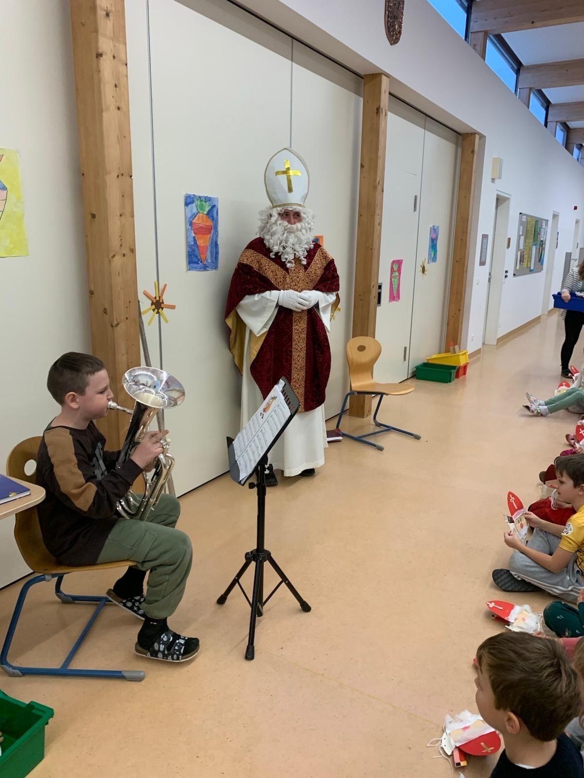Ein Junge spielt die Trompete, während er eine grüne Uniform trägt. Hinter ihm steht ein Mann, der als Weihnachtsmann verkleidet ist und zuschaut. Der Raum ist mit Kindern gefüllt, die auf dem Boden sitzen und die Aufführung beobachten.