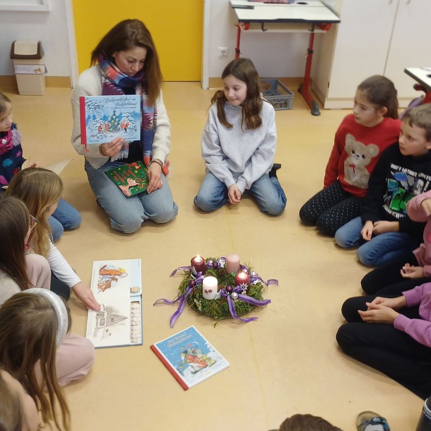 Eine Frau liest Kindern in einem Kreis ein Weihnachtsbuch vor, mit einer Kerzengirlande auf dem Boden. Einige Kinder schauen in Bücher, andere beobachten die Leserin.