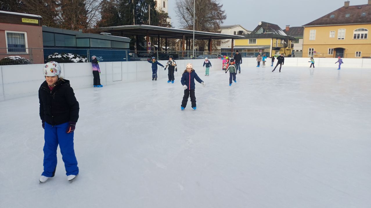 Mehrere Kinder eislaufen auf einer Eisbahn mit einem Gebäude und Bäumen im Hintergrund. Einige tragen Helme.