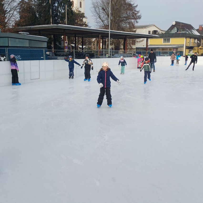 Mehrere Kinder eislaufen auf einer Eisbahn mit einem Gebäude und Bäumen im Hintergrund. Einige tragen Helme.