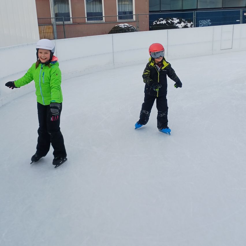 Zwei Kinder, die auf einer Eisbahn Schlittschuh laufen, eines lächelt und das andere trägt einen roten Helm. Sie tragen Jacken und Handschuhe. Dahinter steht ein Gebäude mit einem Zaun.