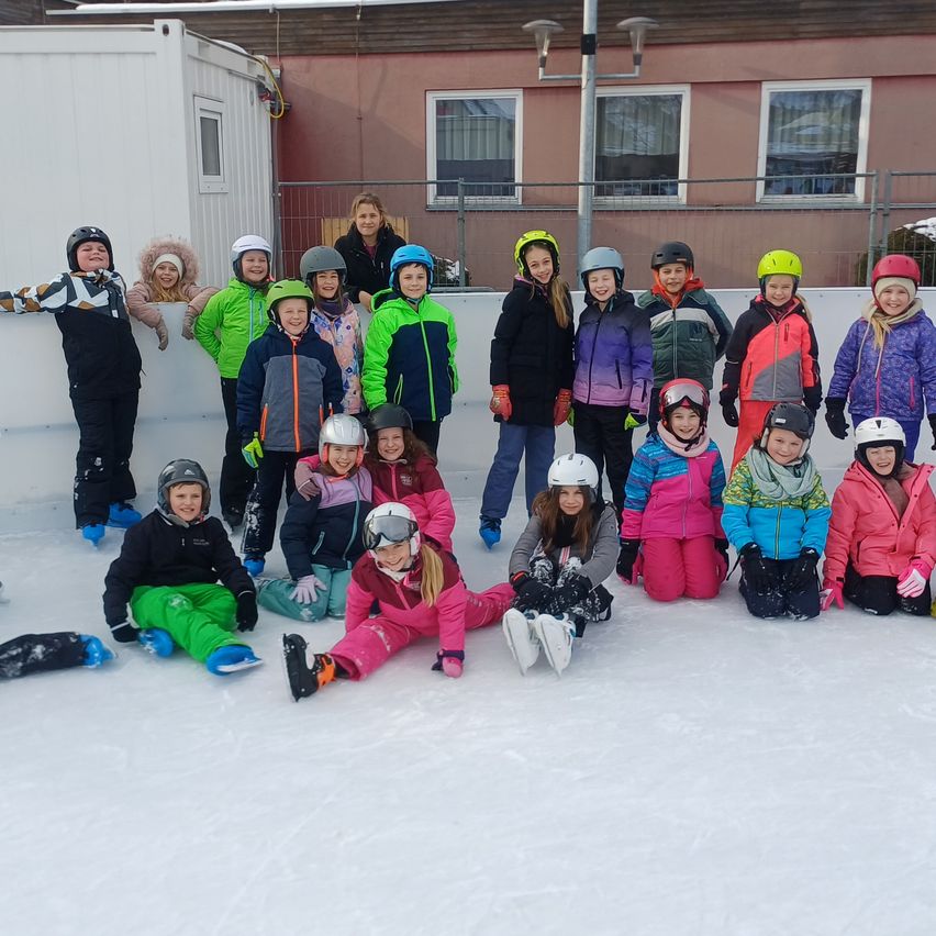 Eine Gruppe von Kindern und Erwachsenen posiert für ein Foto auf einer Eisbahn, sie tragen Winterkleidung und Helme. Hinter ihnen steht ein Gebäude mit einem Zaun und Fenstern.