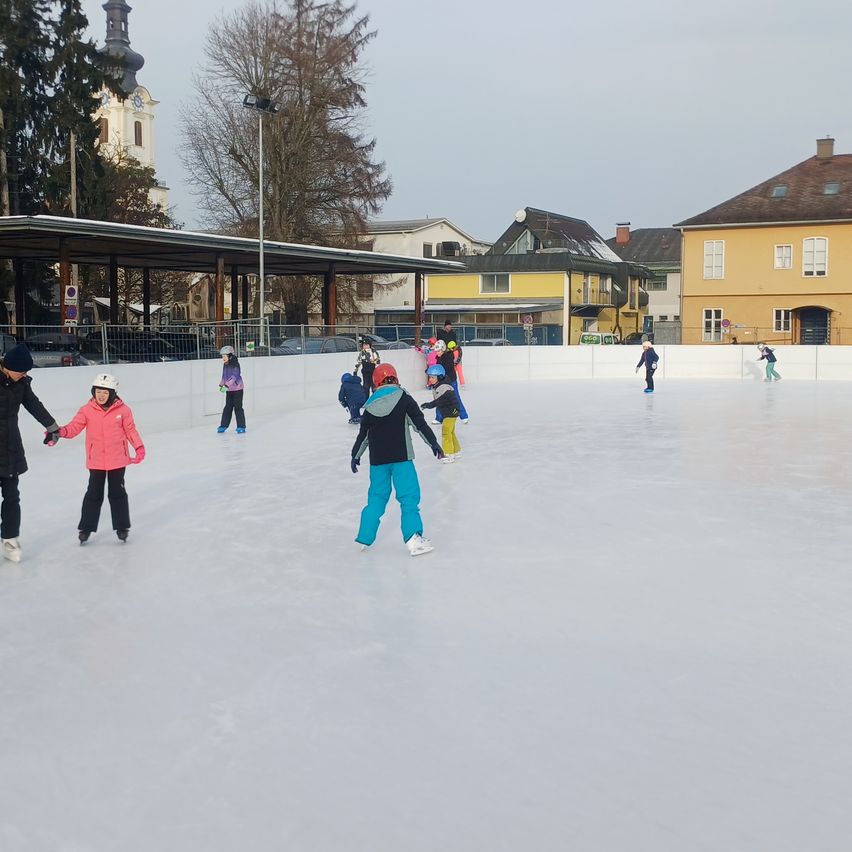 Mehrere Menschen eislaufen auf einer Freiluftbahn. Einige halten sich an den Händen. Ein Gebäude mit braunem Dach steht hinter ihnen.