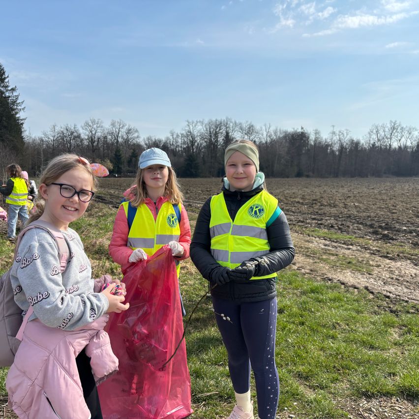 Drei Mädchen in gelben Westen, eines hält einen roten Beutel, posieren auf einer Wiese. Bäume und ein klarer Himmel im Hintergrund.