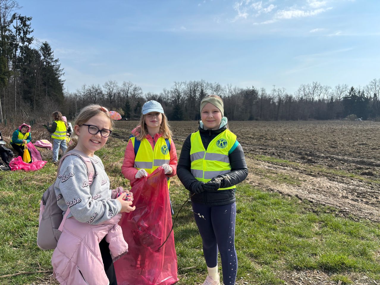 Drei Mädchen in gelben Westen, eines hält einen roten Beutel, posieren auf einer Wiese. Bäume und ein klarer Himmel im Hintergrund.