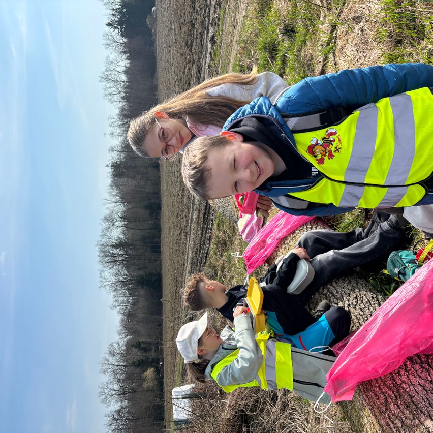 Gruppe von Kindern mit Warnwesten und Mützen ruht sich in einem Feld aus, lächelt, mit rosa Bändern auf dem Boden.