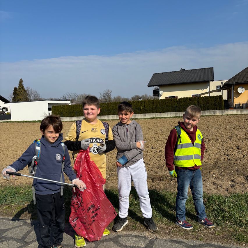 Vier Jungen stehen vor einem Feld, zwei halten einen roten Müllsack. Einer trägt eine gelbe Weste und ein anderer ein gelbes Shirt mit 'ANTIGAM'. Dahinter ist ein Haus unter blauem Himmel zu sehen.