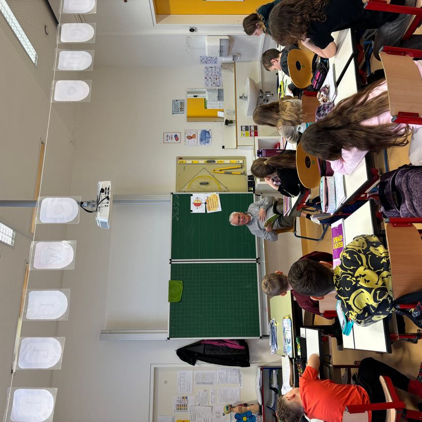 Ein Klassenzimmer mit Schülern, die an Schreibtischen sitzen. Ein Lehrer liest ein Buch und lehnt sich an eine grüne Tafel. Es gibt Papiere und Poster an der Wand.