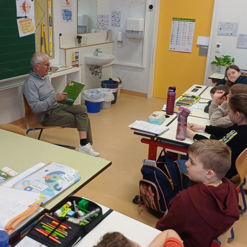 Ein älterer Mann liest ein Buch vor einer Klasse von Kindern. Sie sitzen an Tischen, einige mit Schulsachen. Ein Waschbecken und ein Mülleimer sind im Hintergrund.