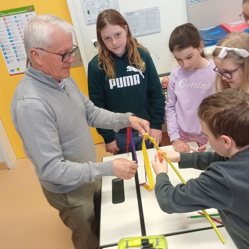 Ein älterer Mann zeigt einer Gruppe von Kindern ein Bastelprojekt mit bunten Bändern. Sie sitzen an einem Tisch in einem Klassenzimmer.