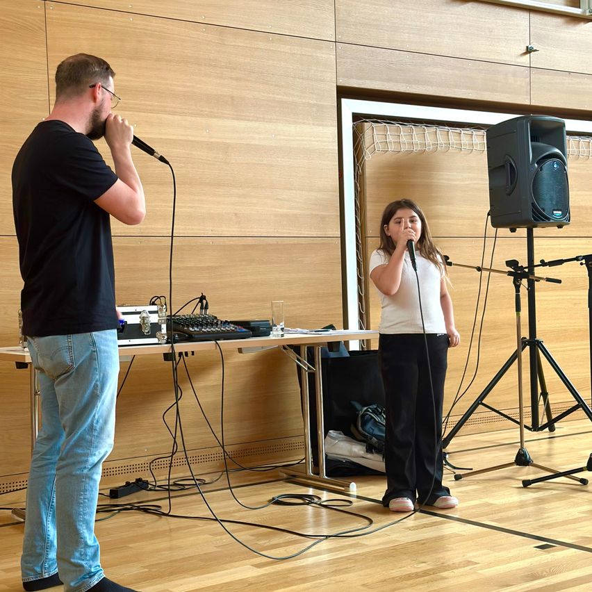 Ein Mann und ein Mädchen singen in Mikrofone auf einem Holzboden, mit einem Tisch und Lautsprecher-Setup hinter ihnen.