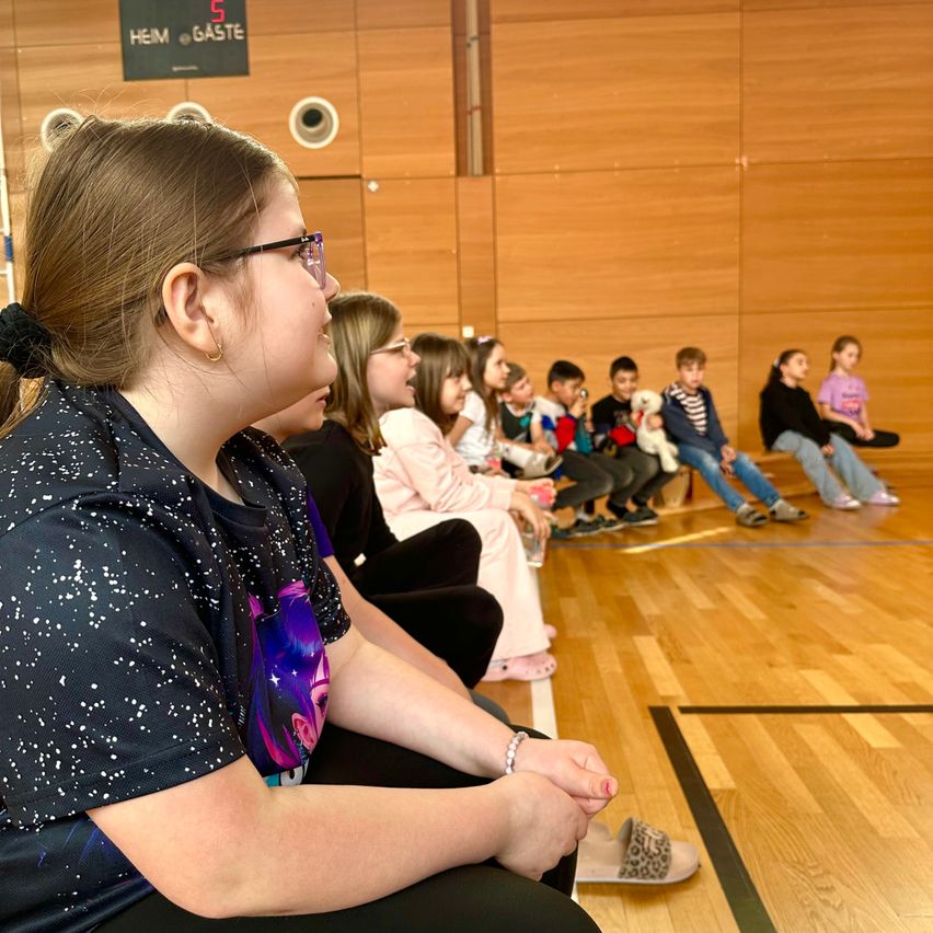 Eine Frau mit einer Brille und einem schwarzen Shirt mit weißen Punkten sitzt auf einer Bank in einem Raum und beobachtet eine Gruppe von Kindern. Sie sitzen in Reihen auf einem Holzboden, einige halten Spielzeug. Eine Wand hinter ihnen hat ein schwarzes Schild mit Text.
