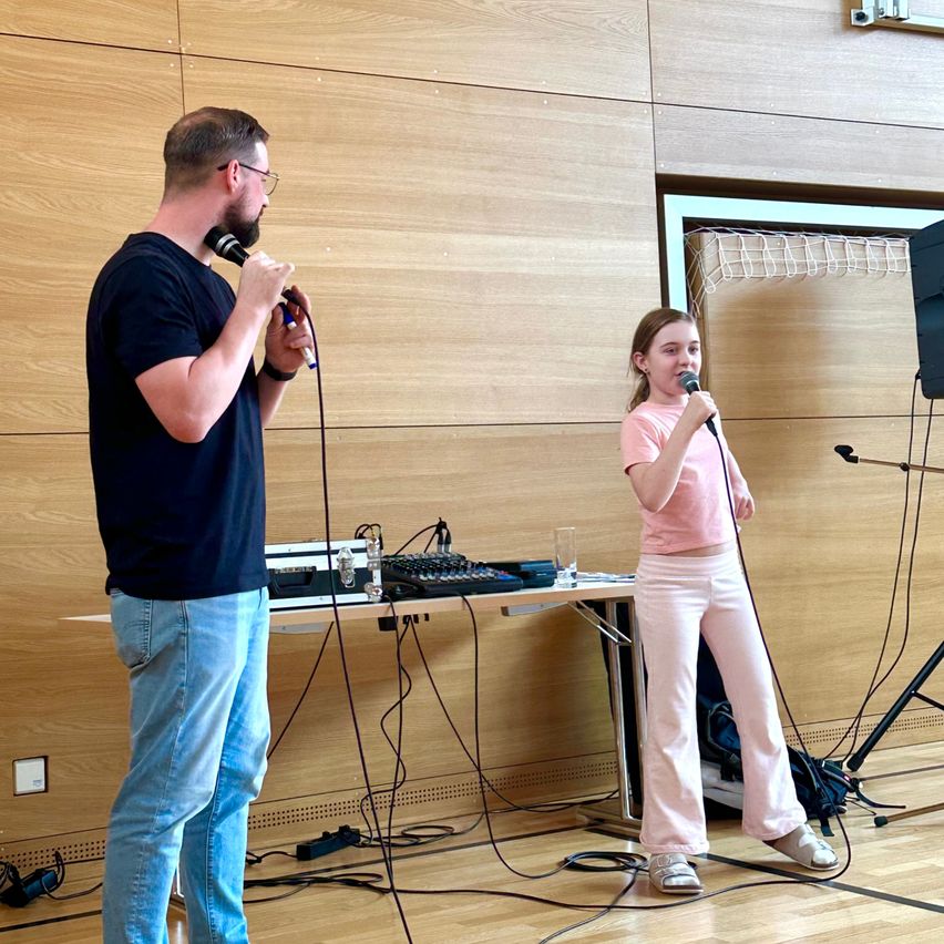 Ein Mann und ein junges Mädchen singen in Mikrofone, wahrscheinlich für eine Aufführung. Hinter ihnen ist eine Holzwand mit einem Tisch, der Tongeräte hält.