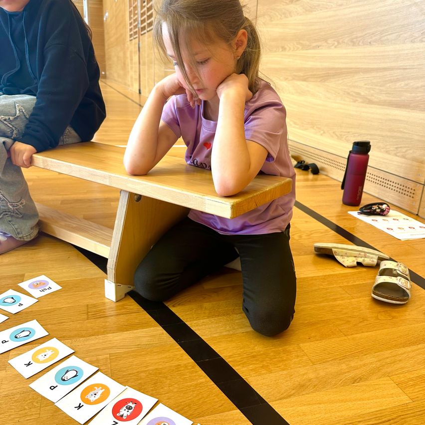 Ein junges Mädchen in einem lila Shirt sitzt auf dem Boden mit den Händen im Gesicht. Sie sitzt vor einem kleinen Holzschreibtisch. Es gibt eine Reihe von Karten auf dem Boden, und eine Wasserflasche und Hausschuhe sind auf der rechten Seite. Eine weitere Person sitzt auf der linken Seite.