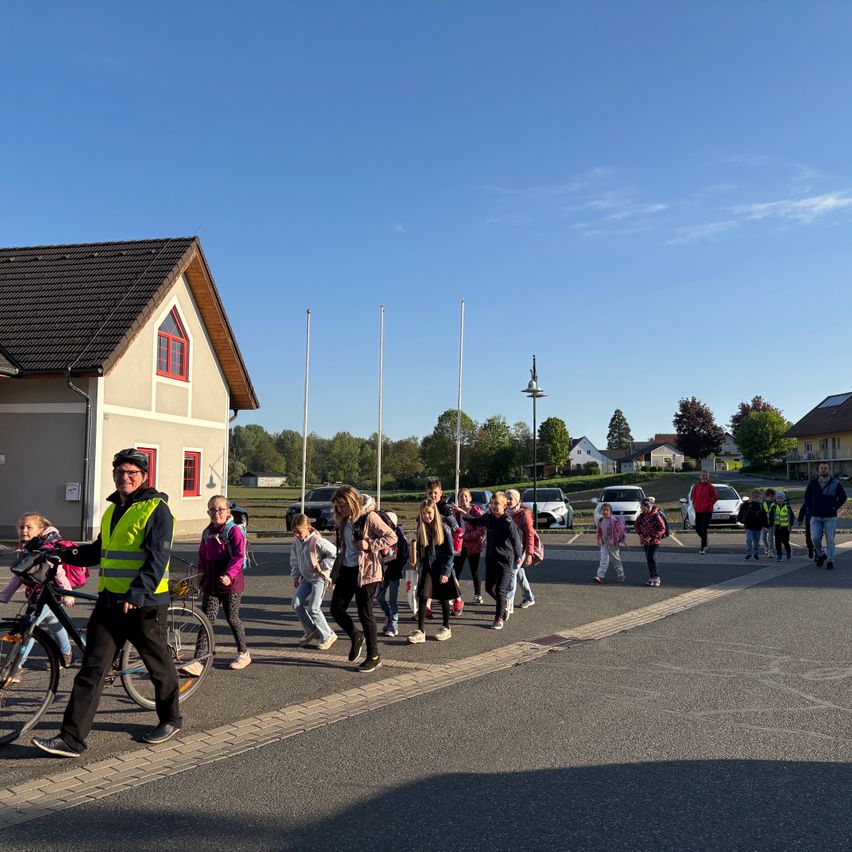 Eine Gruppe von Kindern und Erwachsenen, einschließlich eines Mannes mit einem Fahrrad, überquert die Straße unter der Aufsicht eines Mannes in einer reflektierenden Weste.