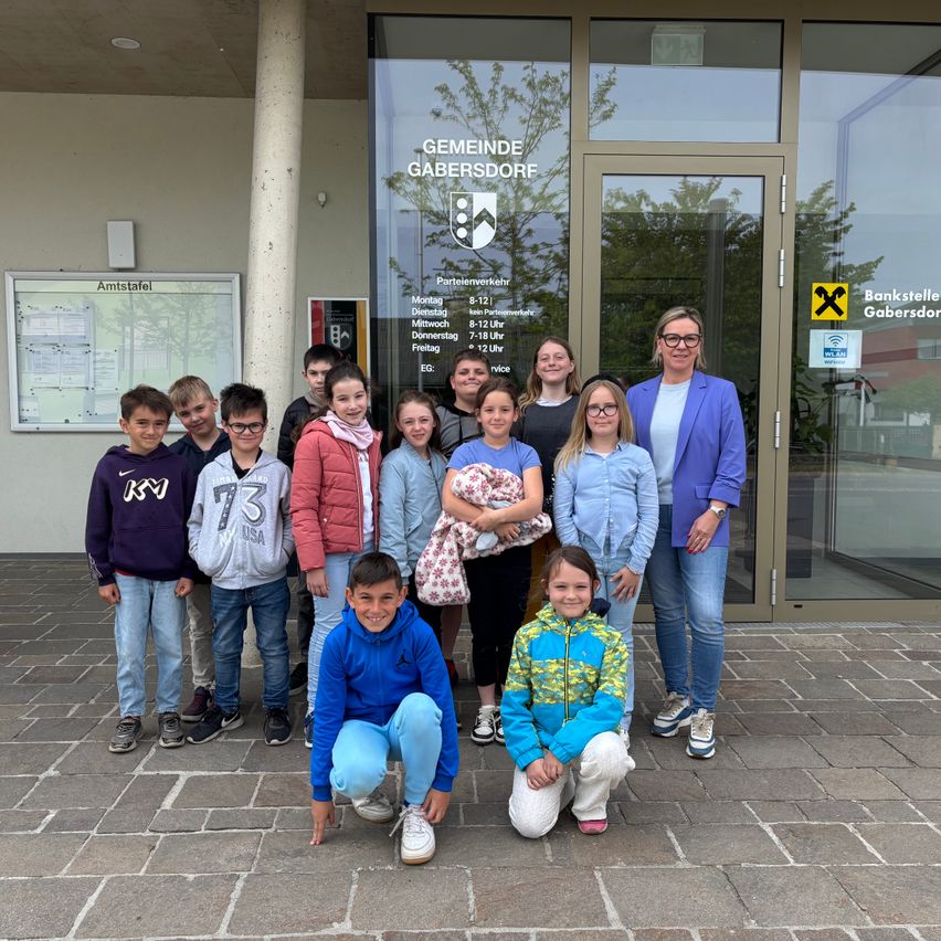 Eine Gruppe von Kindern und zwei Erwachsenen steht vor einem Gebäude, das Gemeinde Gabersdorf heißt, mit einer lächelnden erwachsenen Frau, die ein Baby hält.