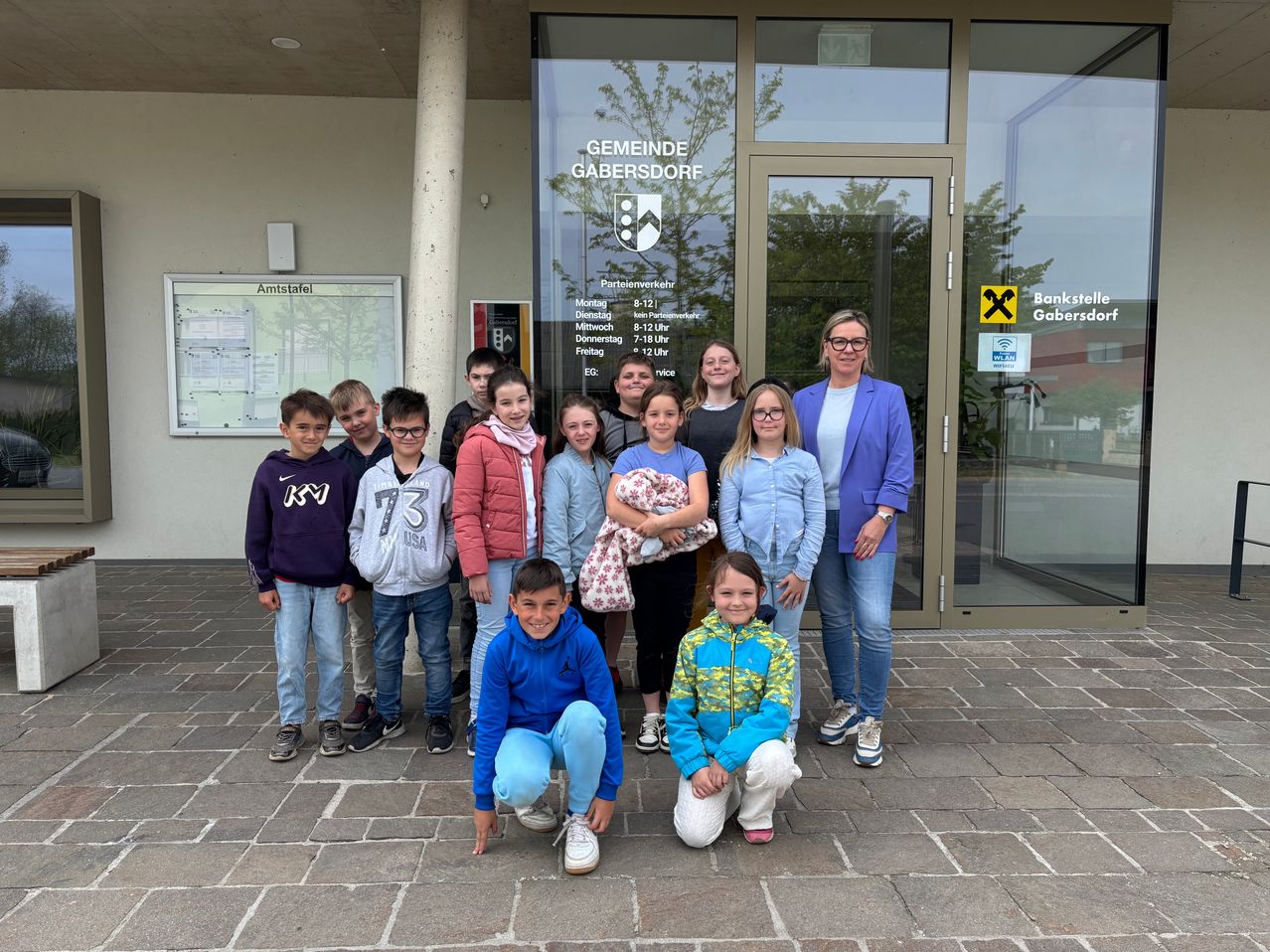 A group of children and two adults stand in front of a building labeled Gemeinde Gabersdorf, with a smiling adult woman holding a baby.
