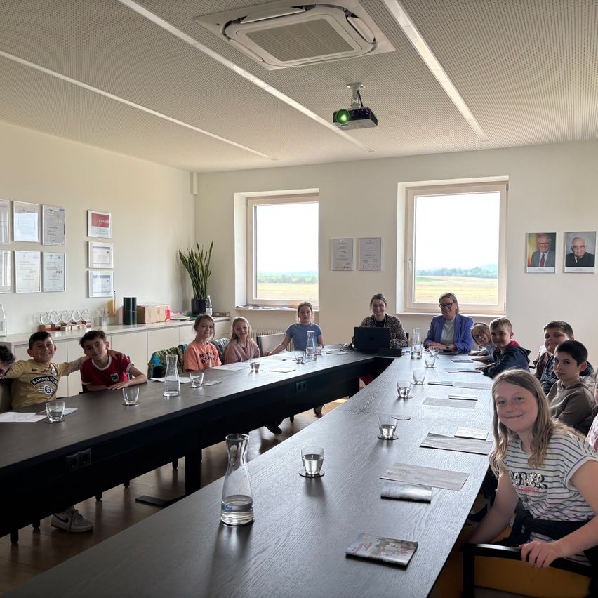 A group of children and adults sit around a long table in a meeting room. The room has large windows, a projector, and framed pictures on the wall.